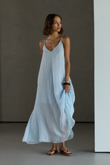 A woman modeling a light blue sleeveless maxi dress with thin straps and a V-neckline, standing in a minimalist studio with a dark gray background.